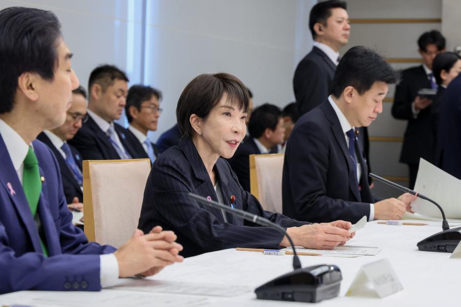 Sanae Takaichi in dark suit jacket sits at a table with microphones between two men, and more men sitting in the room behind her