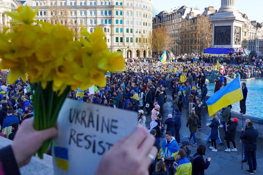 A crowd gathered, holding blue and yellow flags; hands in the foreground hold a sign that says "Ukraine Resist"
