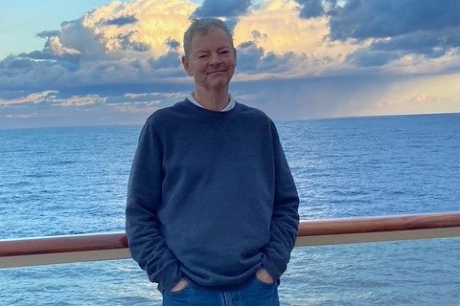 Robert Sullivan, with short gray hair and a closed-mouth smile, leaning against a ship's rail with the sea and clouds behind him.