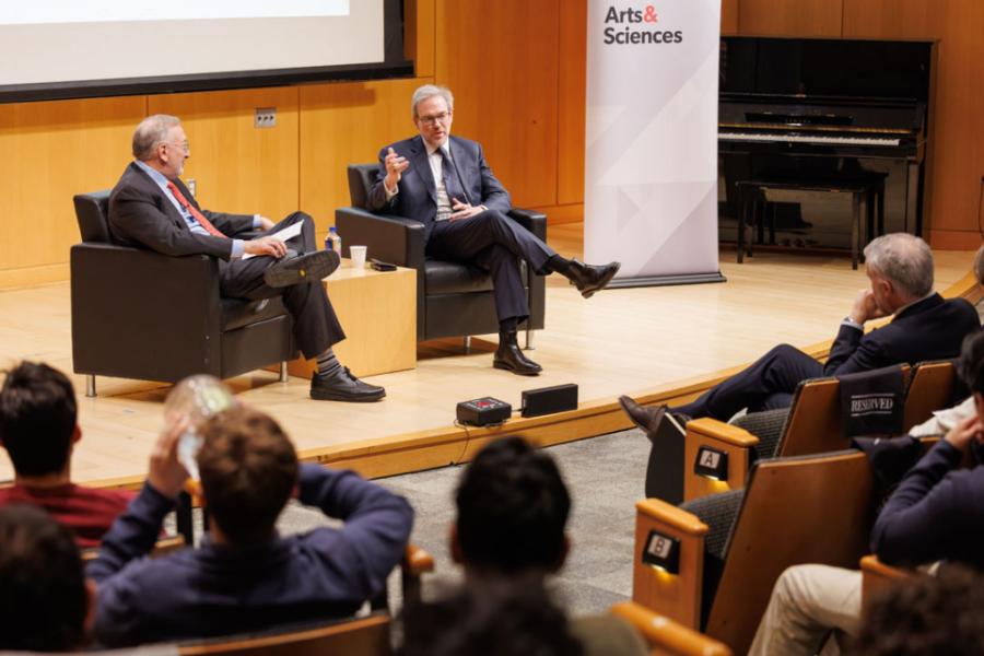 Two people seated in large chairs, conversing in front of an audience