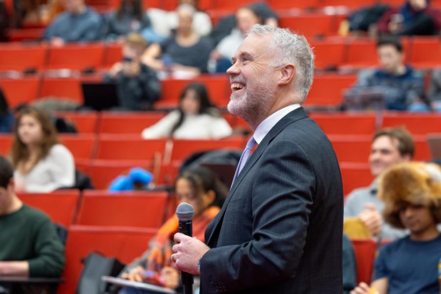 Person in suit holding microphone, in front of a bank of red seats, some occupied