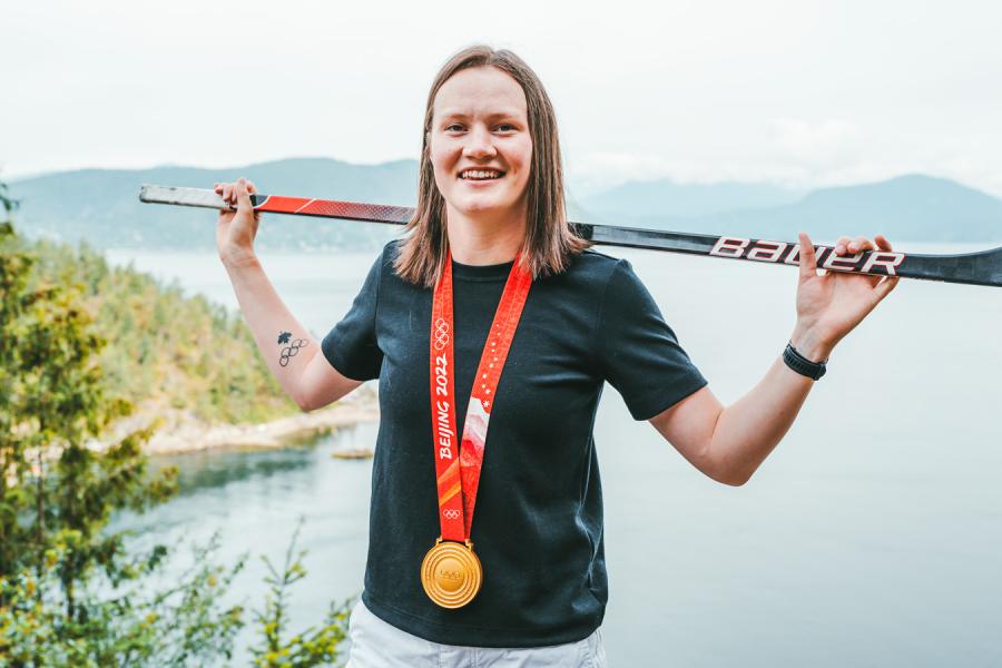 Person standing in front of a body of water, balancing a hockey stick across her back and wearing a gold medal on a red ribbon