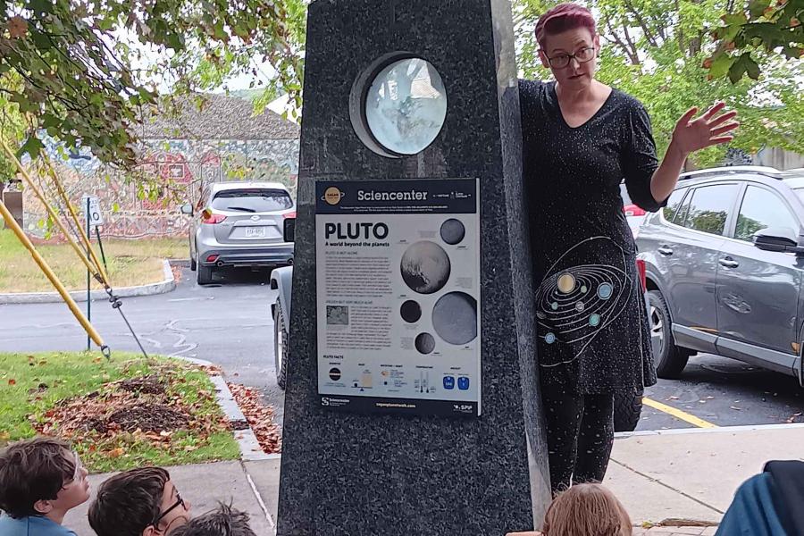 Zoe Learner Ponterio, wearing a dress with a picture of the solar system, stands next to the Sagan Walk Pluto marker, talking to a bunch of kids sitting on the ground looking up at her.