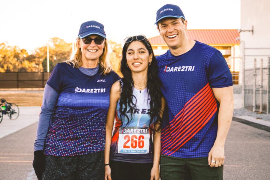 A smiling woman and man in caps and "DARE2TRI" T-shirt stand on either side of a young woman athlete wearing the number "266" on her shirt