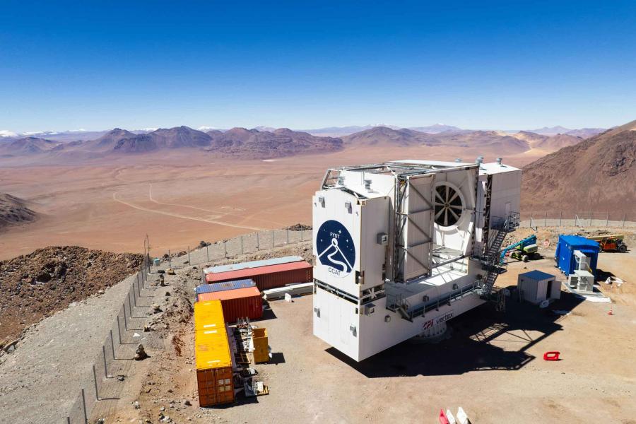 The telescope, a large white structure with the logo on the front, with construction boxes around it and mountain tops and a plateau in the distance.