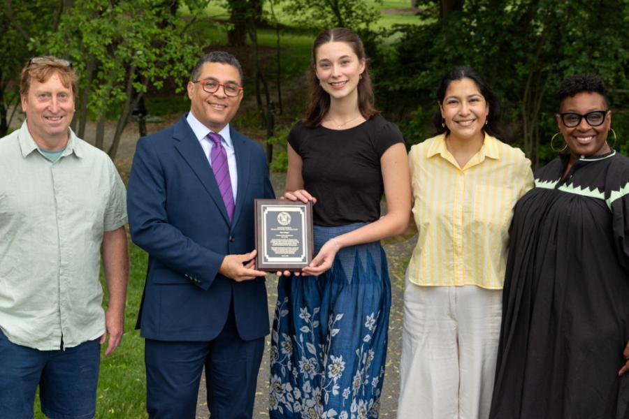 Five people lined up for a photo: in the center a college student award recipient holds a plaque