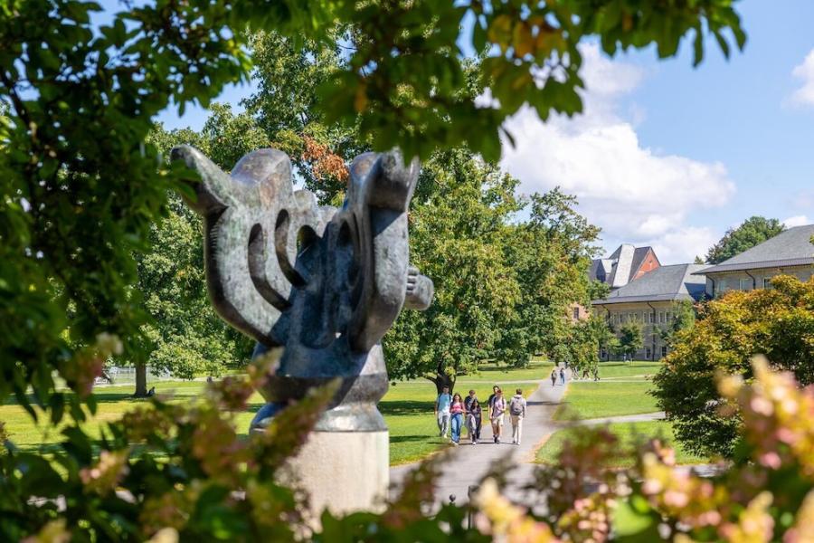 an abstract sculpture in the foreground with a grassy college quadrangle in the distance, bordered by low buildings