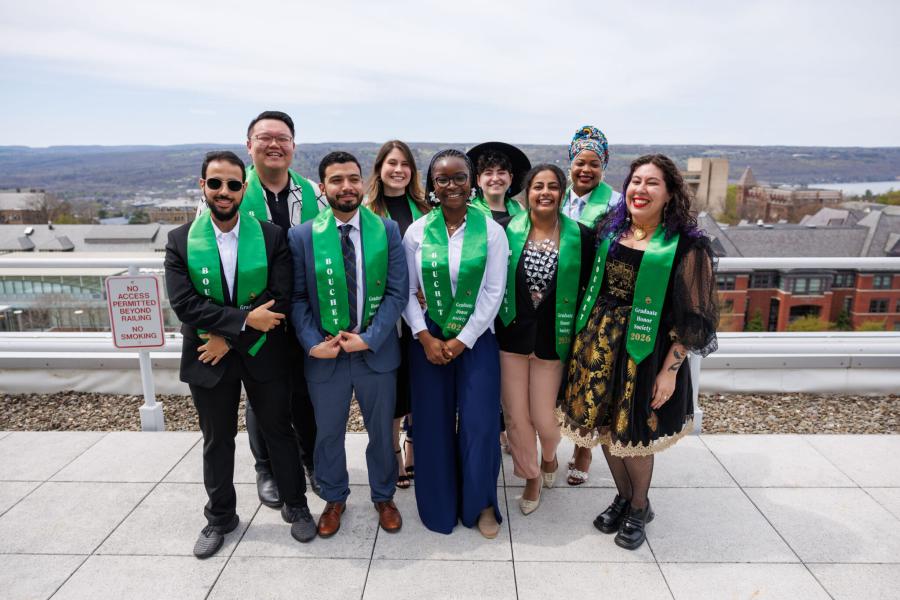 Several people, all wearing green honorary stoles, stand together on a rooftop with a view of a college campus