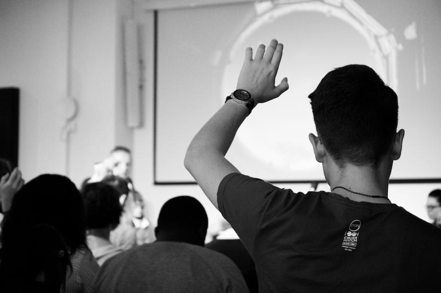 Person seen from the back with hand raised in an audience facing a speaker