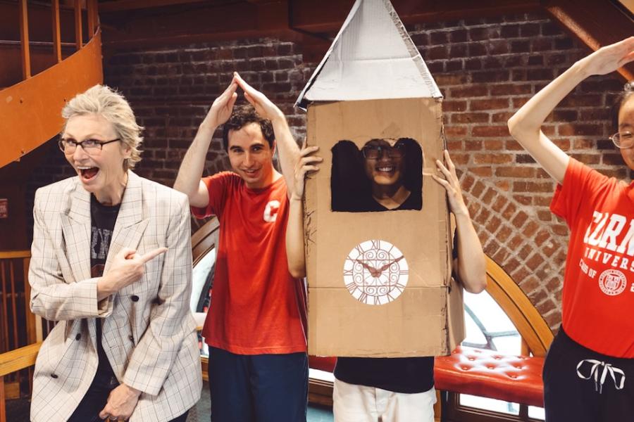 Four people goofing off in a brick-walled room. One wears a cardboard clock tower costume