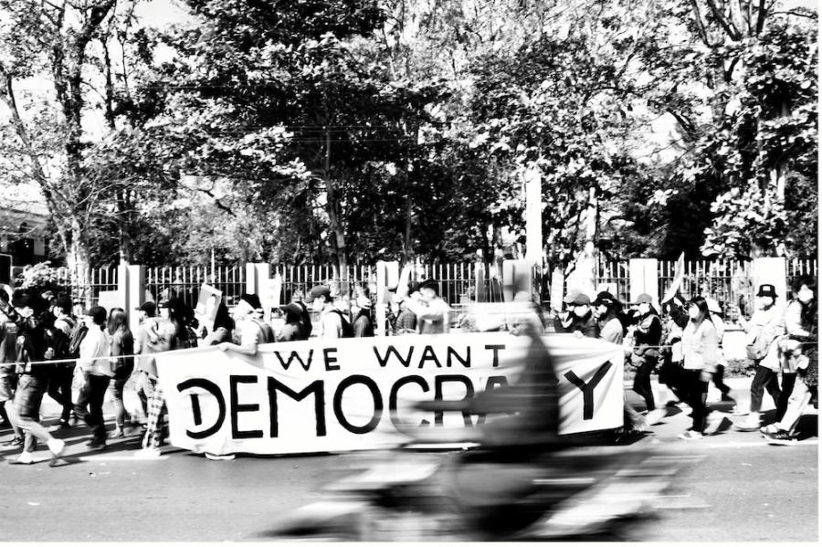 Black and white photo of many people walking together down a street, holding a large banner that reads "We Want Democracy"