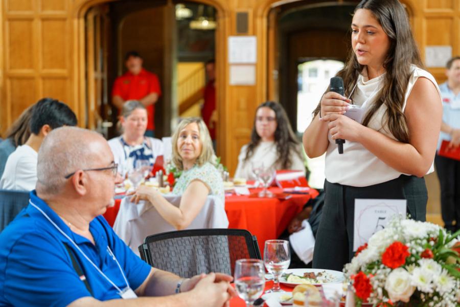 Person speaks into a microphone, directing attention to one other person in a hall full of people sitting at tables 