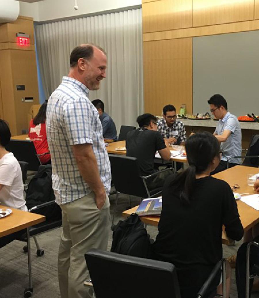  Teacher stands near students, who are writing at a table