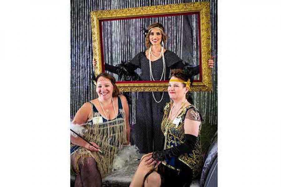 Three women dressed in 1920's dresses with fringes and pearls pose in a giant picture frame