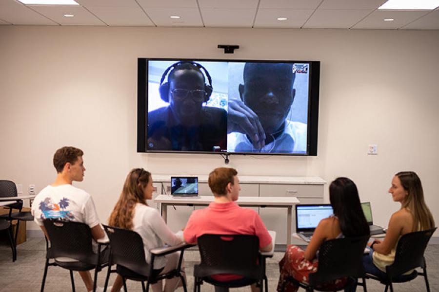 Human to Human team members in a classroom facing a large videoscreen talking to Beta project partners