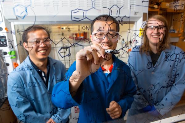 Three people in lab coats behind a clear wall inscribed with organic chemistry symbols