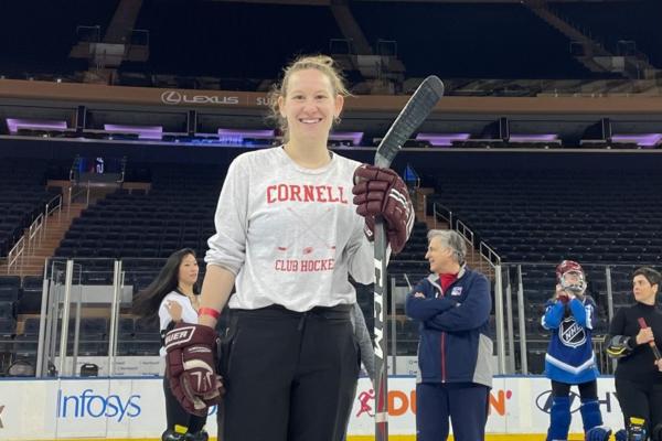 Person wearing hockey gloves and a Cornell t-shirt, carrying a hockey stick and smiling