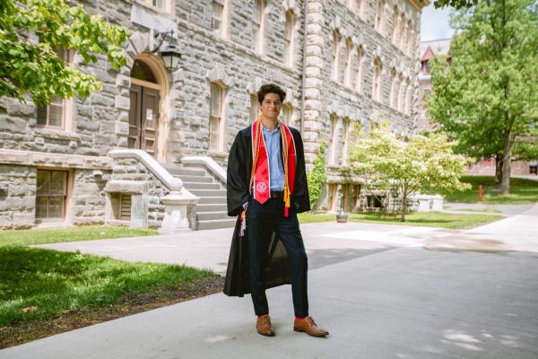 Smiling photo of smiling man with Cornell graduation gown in front of academic builing.