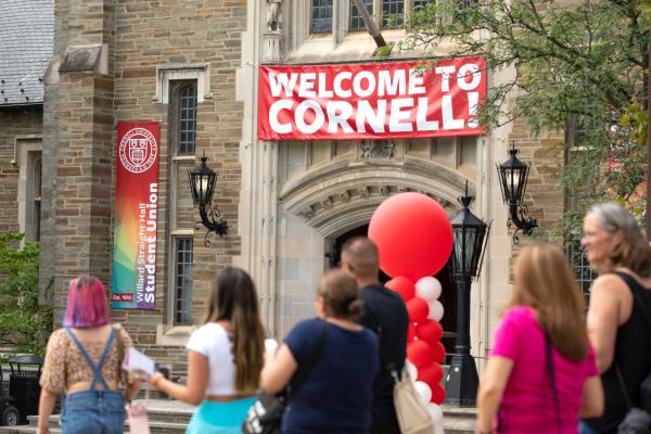 Several people walk past a building with a red and white banner that says "Welcome to Cornell." There are red balloons