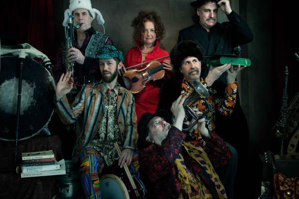 Six people in colorful, odd clothing, holding and playing musical instruments including fiddle, trumpet and saxophone