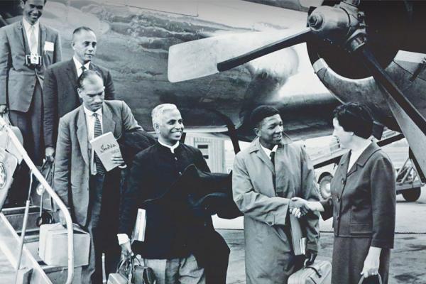 Black and white historic photo: Five people getting off a plane; one is shaking hands with a person wearing a suit at the bottom of the stairacase