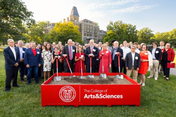 A crowd of about 75 people stands behind a low box full of dirt; six people in the front hold shovels with red handles during a ceremonial "groundbreaking" event