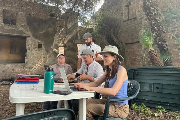 Four people work at a plastic patio table in the midst of ancient ruins: they are archaeologists on an excavation site