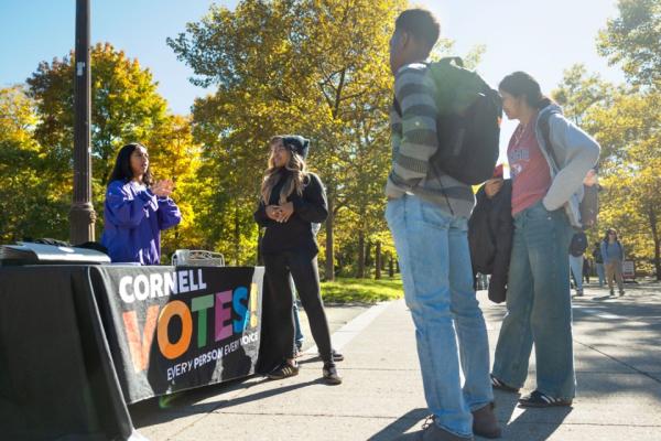 People with backpacks and jeans stand in front of a table, set outdoors, that's labeled "Cornell VOTES"