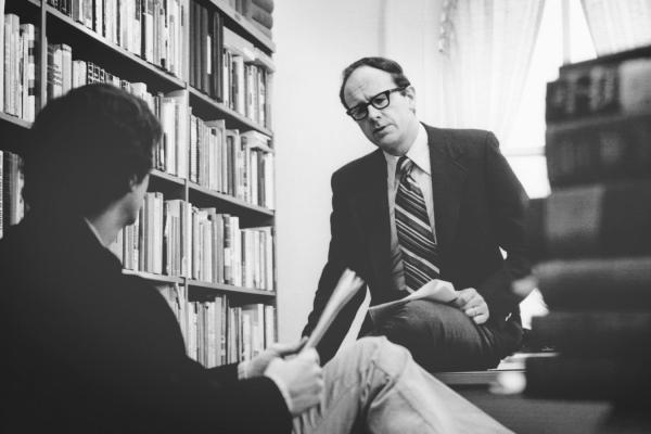 Walter LaFeber in suit and striped tie in his office with bookshelf on one side and a pile of books on the other, talking to someone.