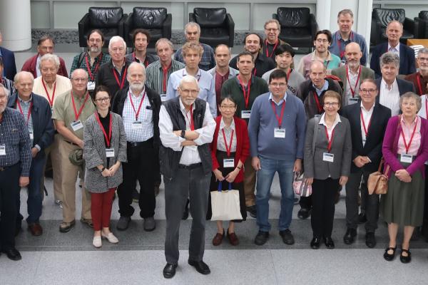 Peter Lepage, wearing a black vest, stands in front of a large group of people assembled to talk about physics and honor his work