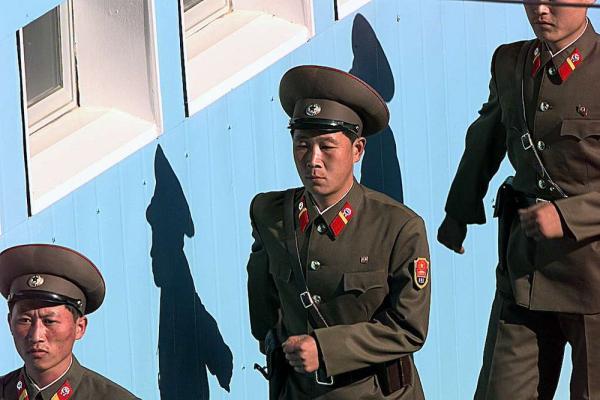 Soldiers in brown formal uniforms march in line beside a blue wall