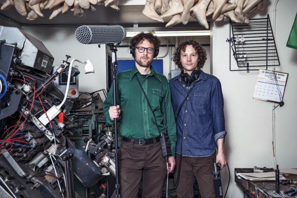 Two people in casual clothes stand in a room full of bric-a-brac, holding professional grade recording equipment