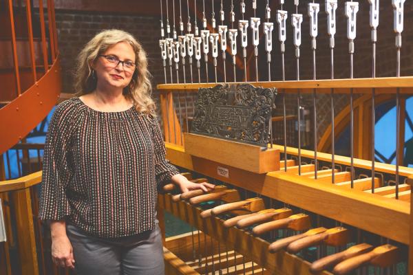 Jennifer Lory-Moran, in multi-colored stripped shirt and long blond hair, is smiling while standing next to the controls for the Cornell chimes up in McGraw Tower.