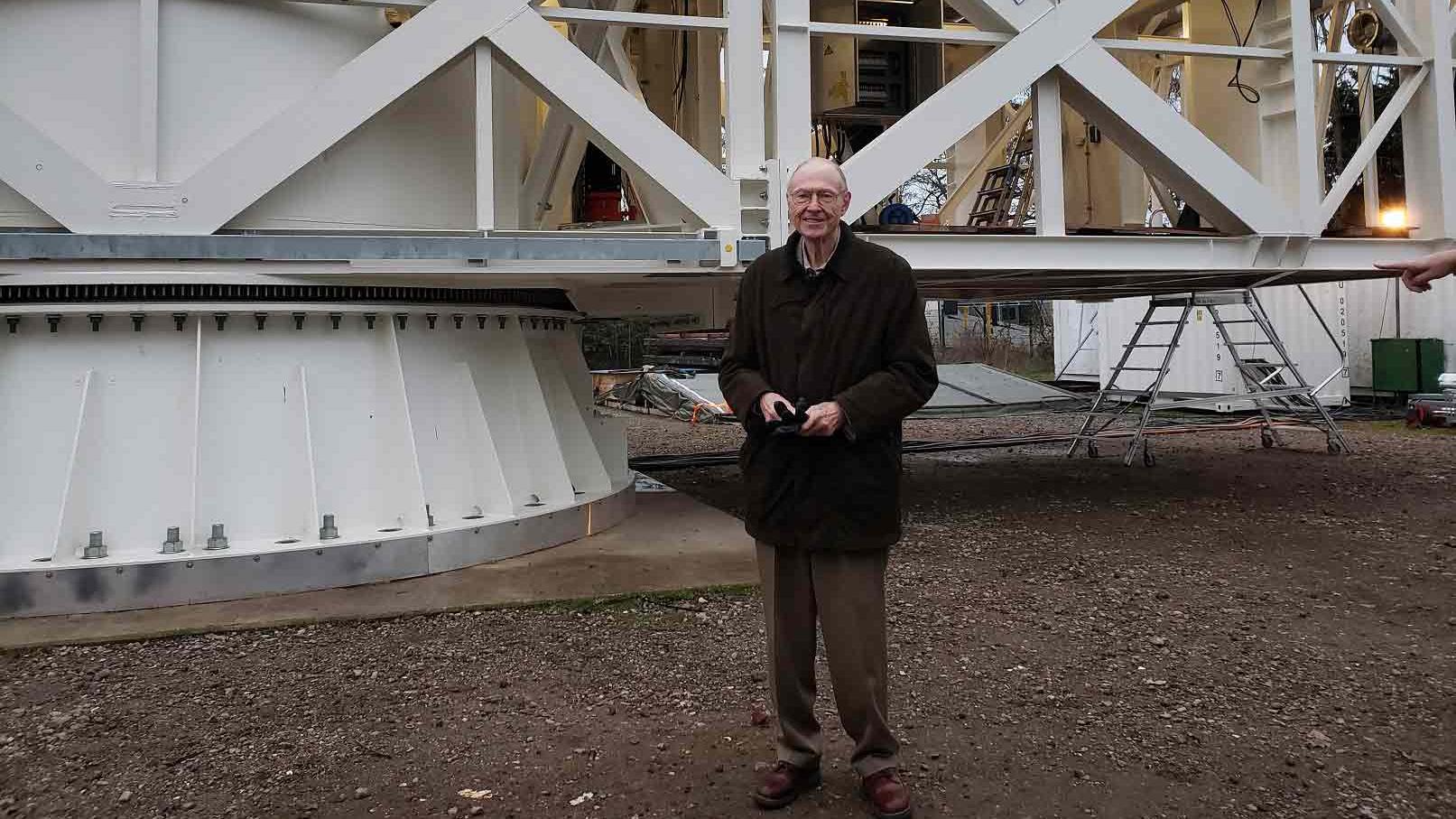 Fred Young, in glasses and black coat, standing in front of giant white metal structure with lattice beams and round base.