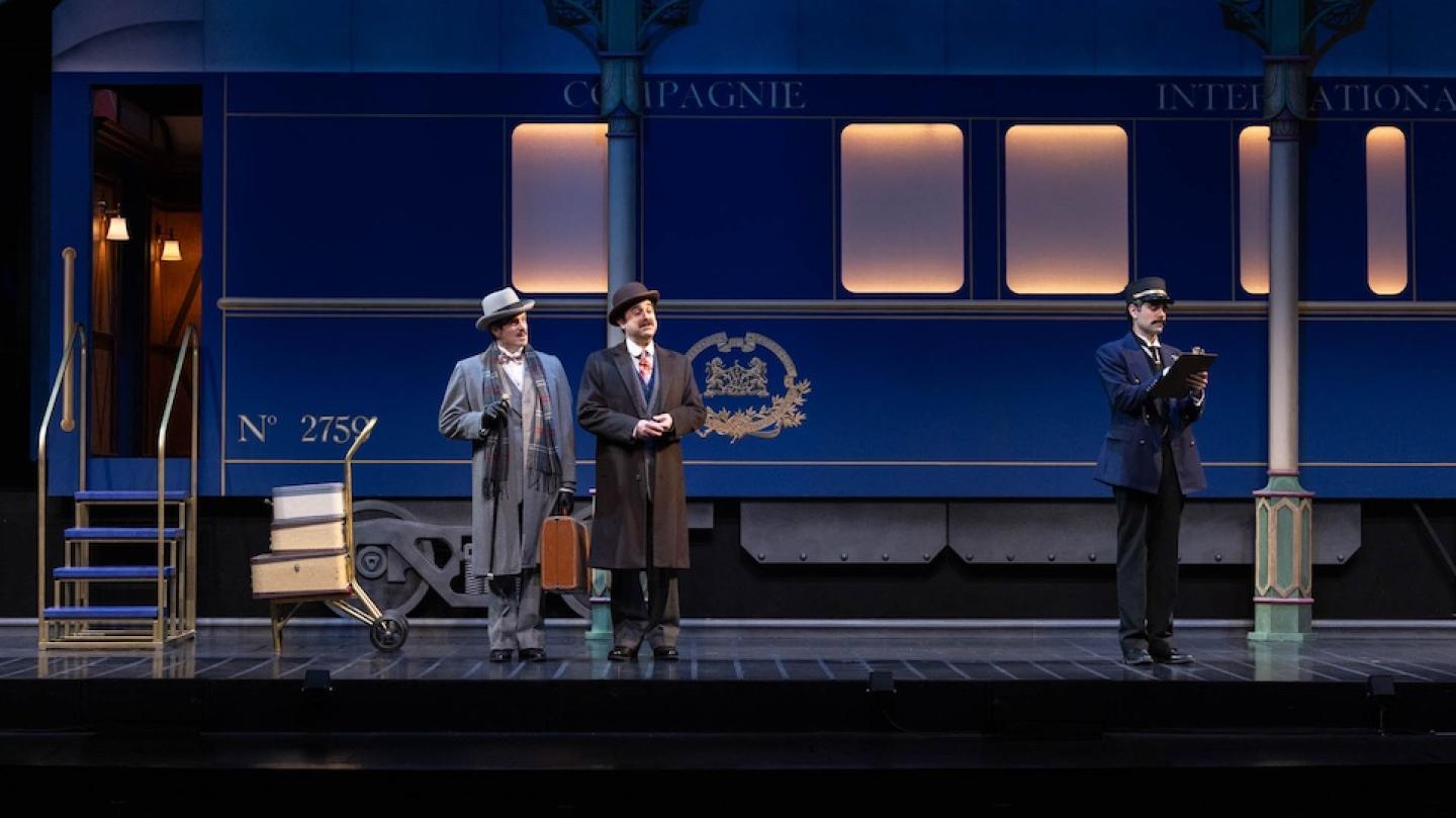 Three people, actors in a play, stand in front of a set that looks like a train car