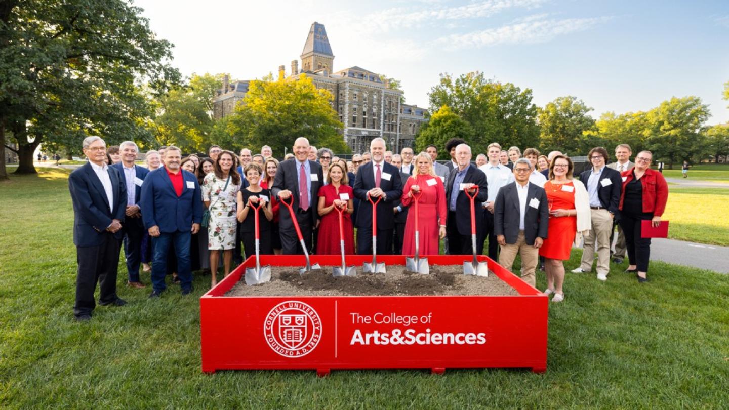 A crowd of about 75 people stands behind a low box full of dirt; six people in the front hold shovels with red handles during a ceremonial "groundbreaking" event