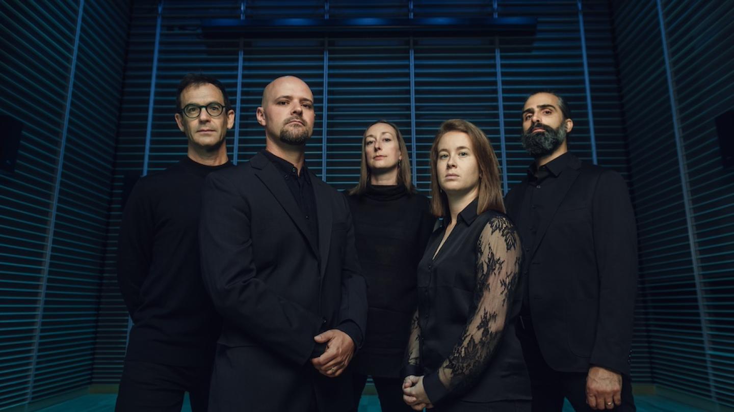 Five people wearing black pose against a blue and black background. They look forboding.