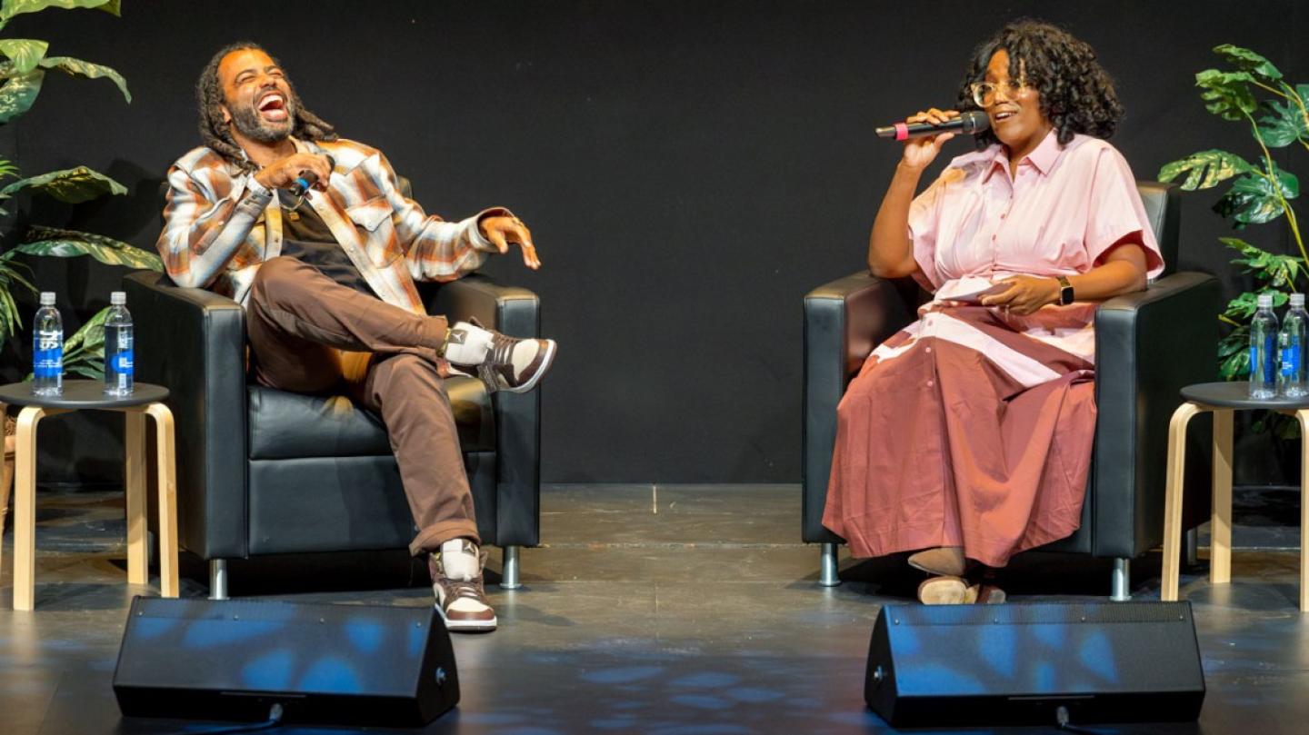 Two people on a stage, sitting in arm chairs, holding microphones