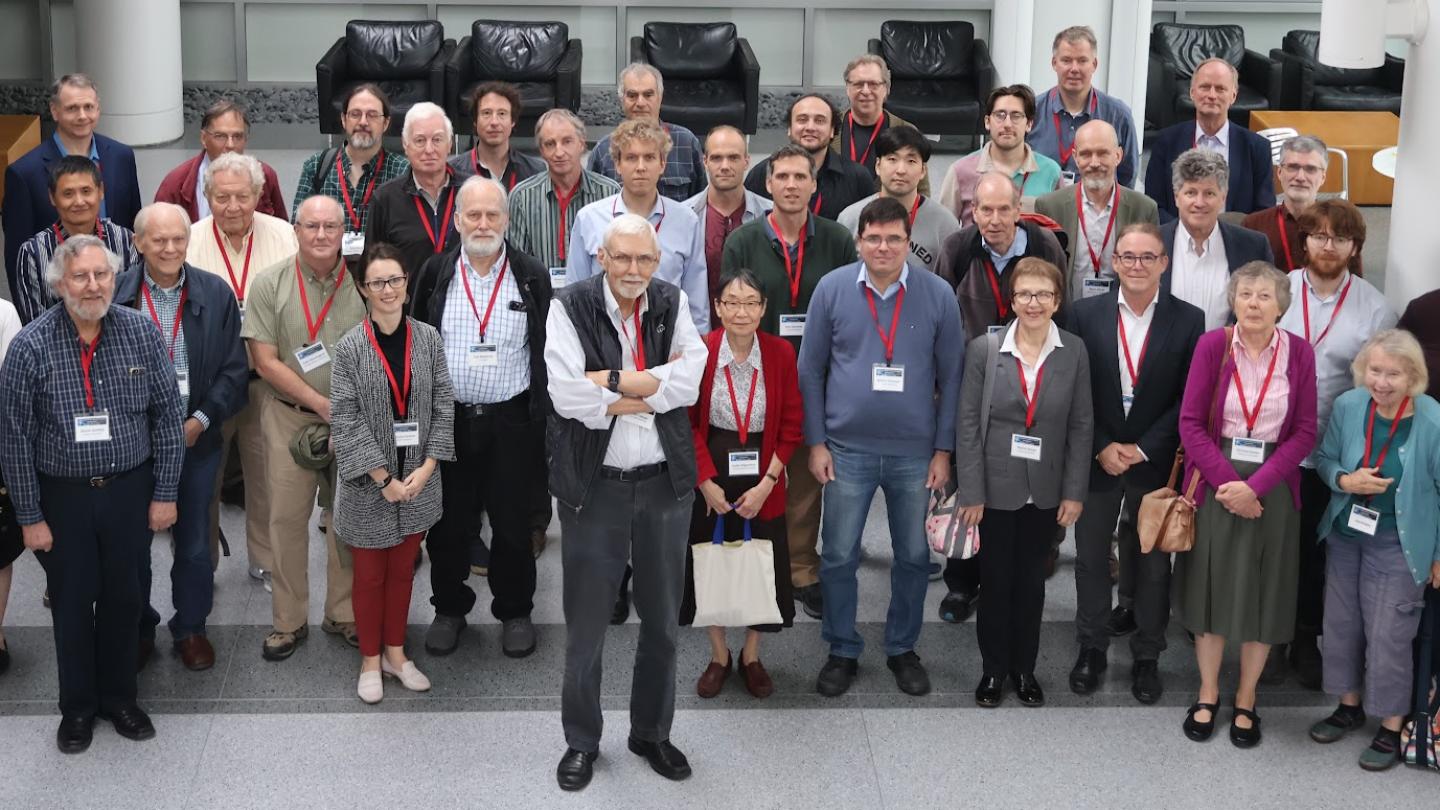 Peter Lepage, wearing a black vest, stands in front of a large group of people assembled to talk about physics and honor his work