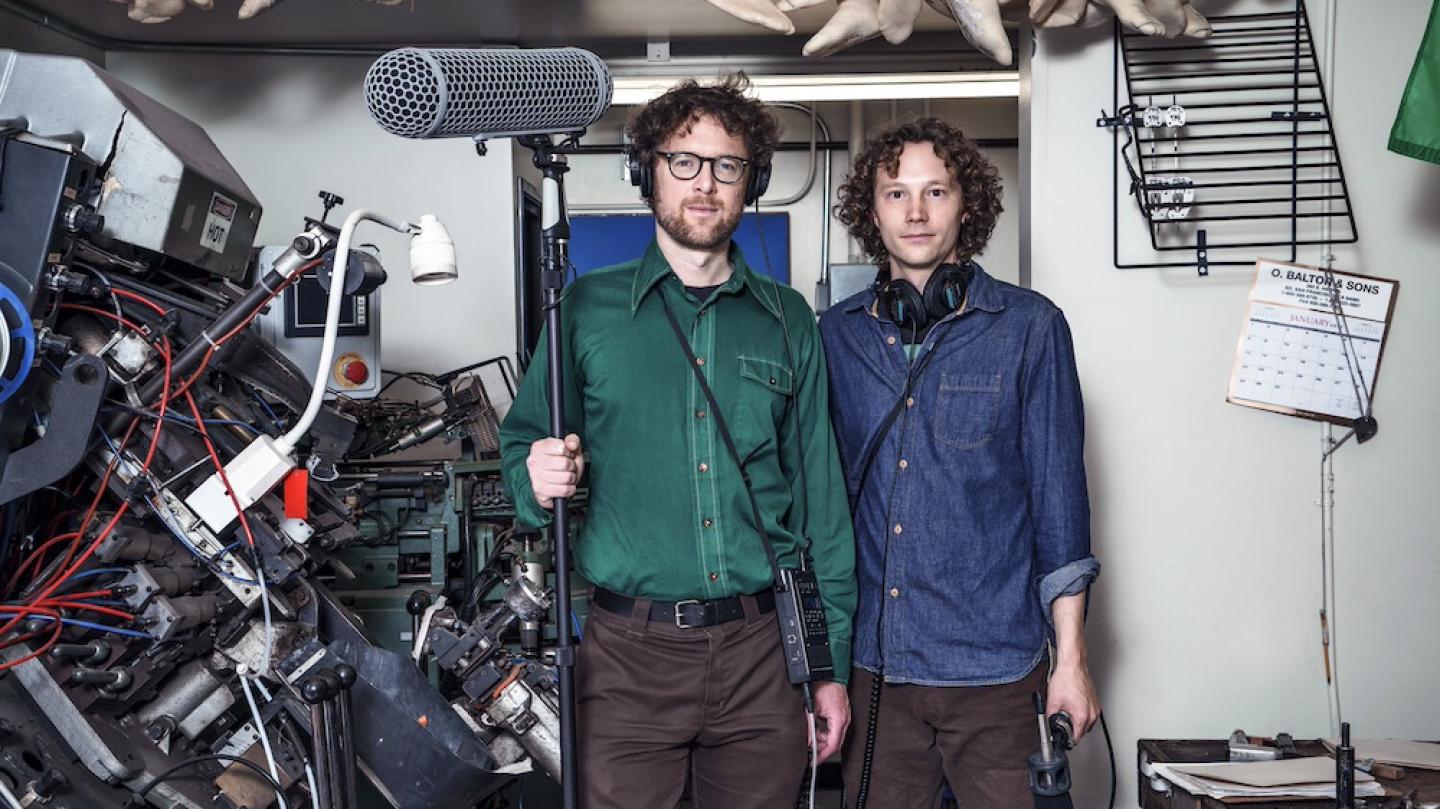 Two people in casual clothes stand in a room full of bric-a-brac, holding professional grade recording equipment