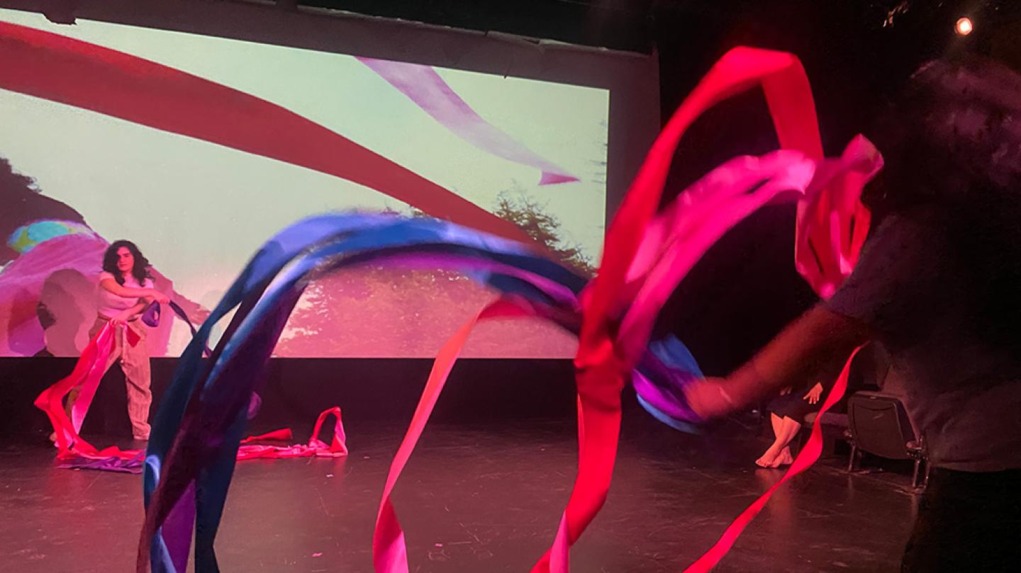 Two colorful ribbons in motion fill the frame, with a dancer in the background, far left