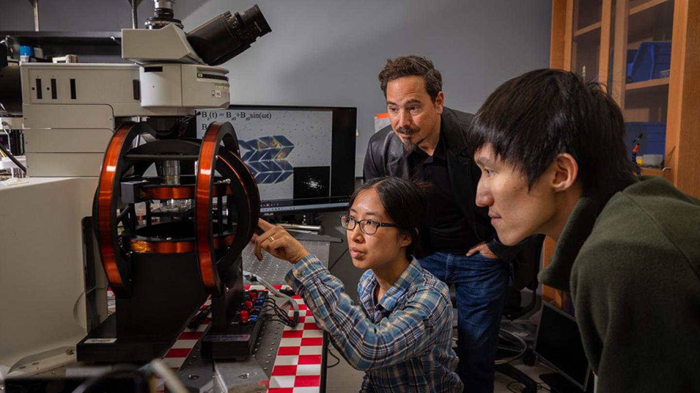 Three people look intensely at a small black and red machine in a science laboratory