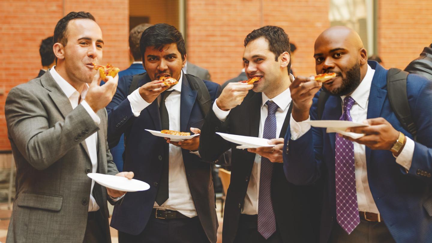 Four people wearing suits, all chomping on slices of pizza and holding plates