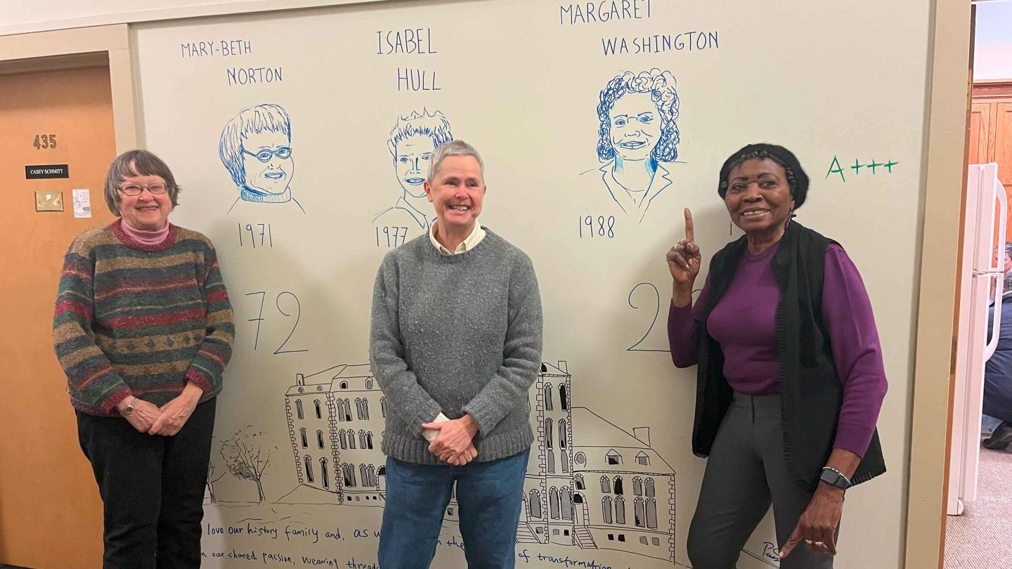 Mary Beth Norton, Isabel Hull and Margaret Washington, all smiling, standing in front of the drawings of their faces on the wall