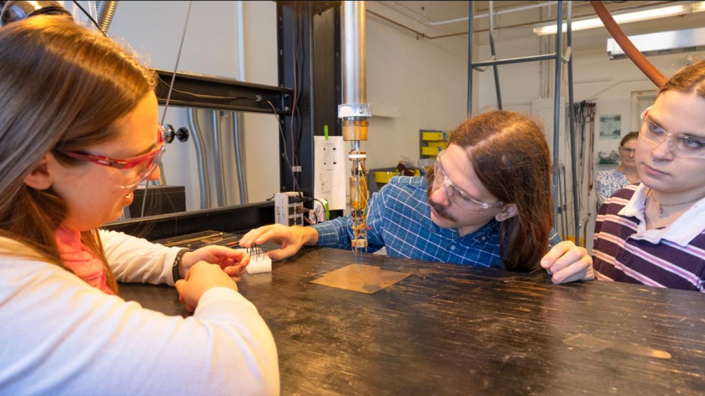 Three people wearing lab safety glasses look at a small scientific device