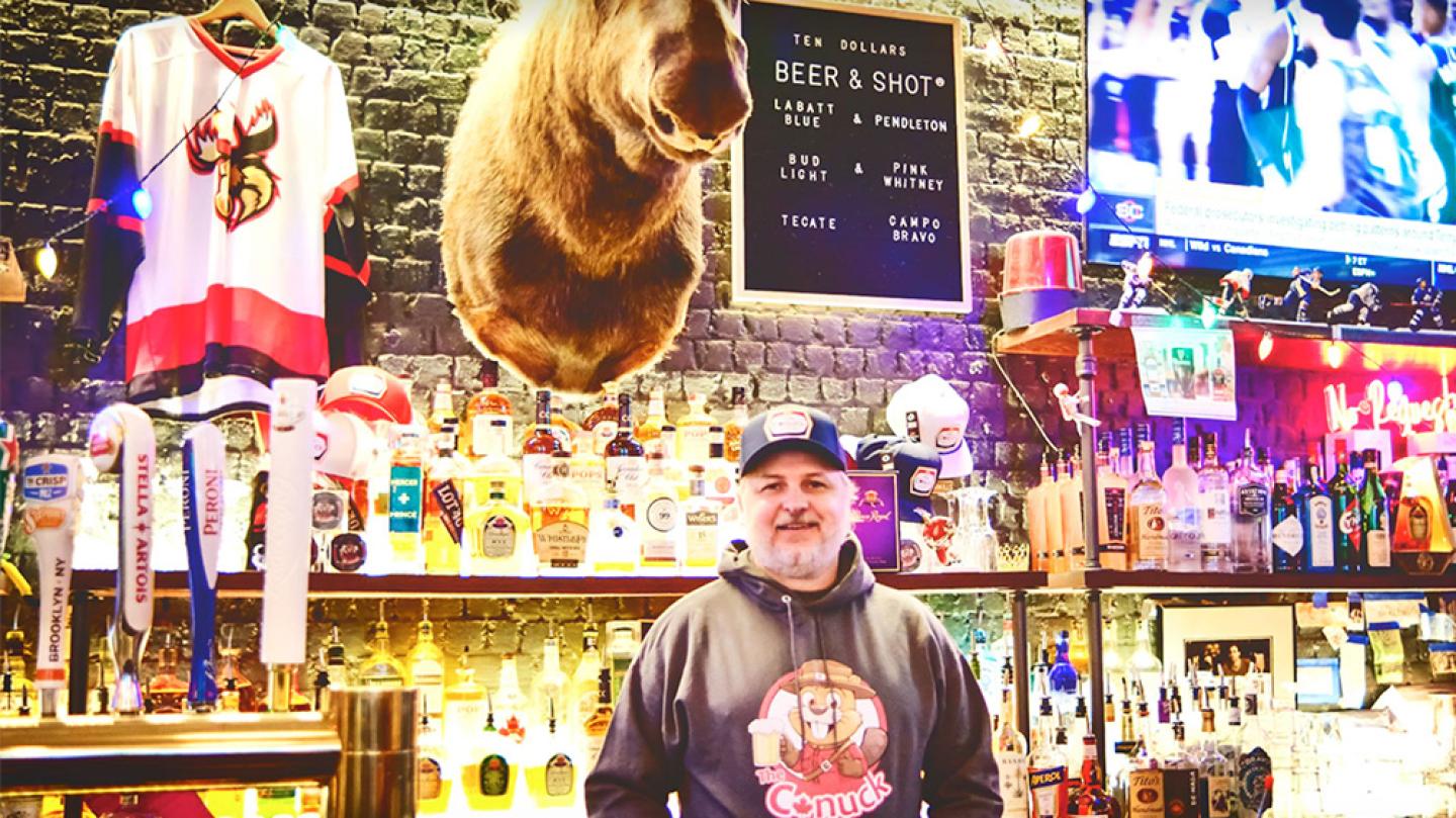 Person standing behind a bar decorated with a hockey jersey, a beer & shot list and a mounted moose head. Lots of neon lights and two illuminated shelves of liquor bottles