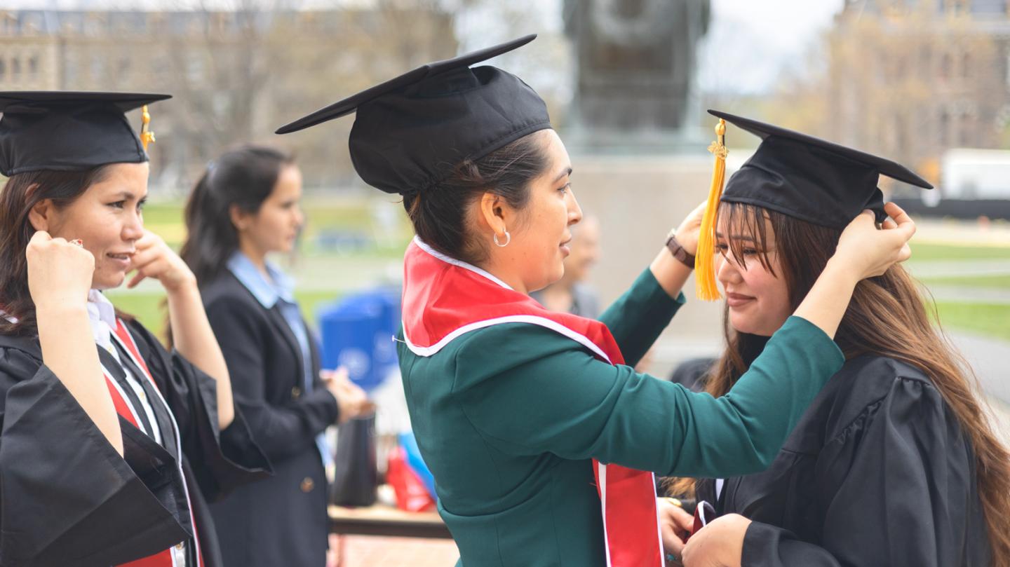 One person wearing graduation cap helps another with hers