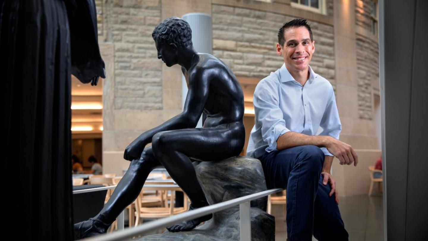 Person sitting casually back to back with a statue of a seated human figure carved from black stone