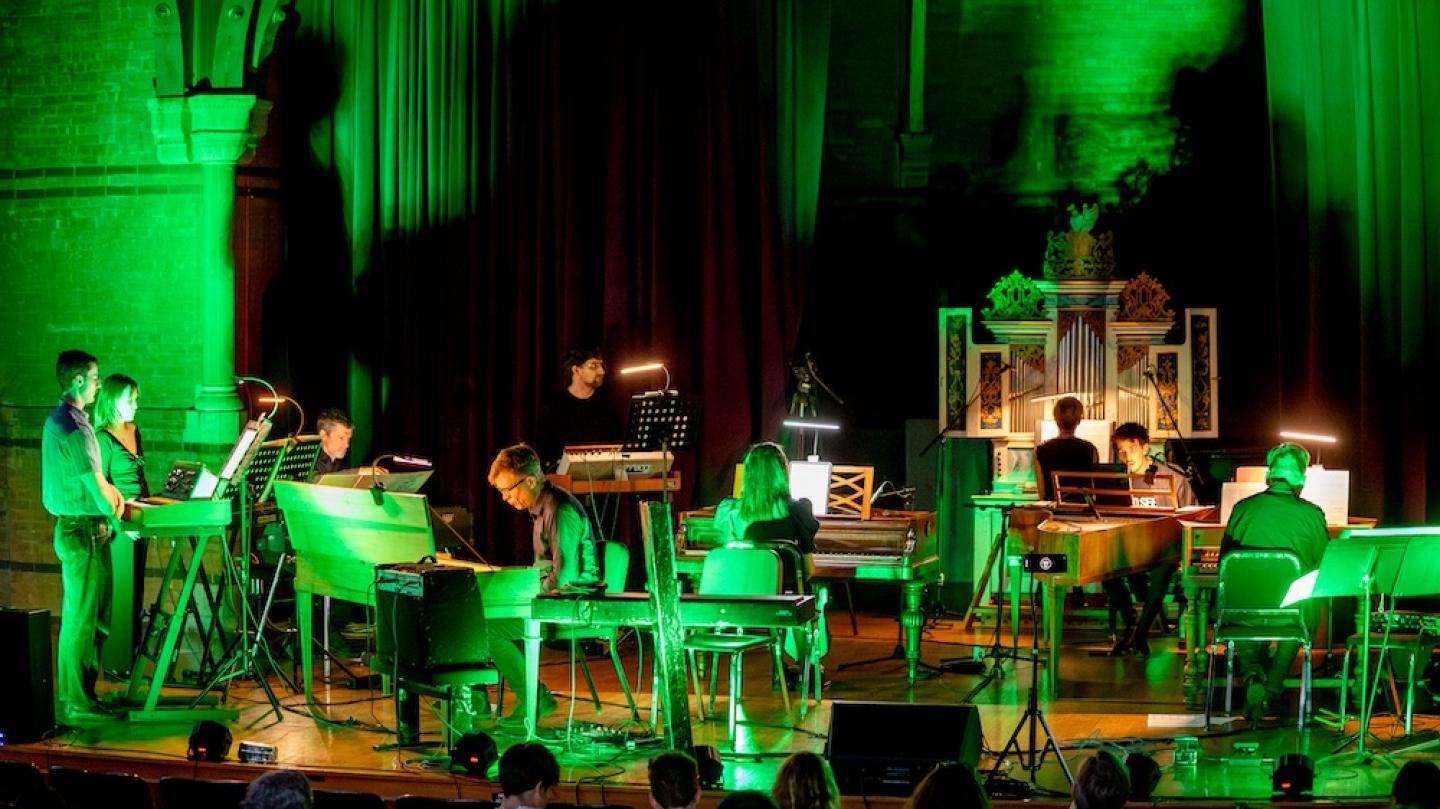 A stage bathed in green light: several people play on keyboard instruments, including standing sythesizers, pianos, and an organ