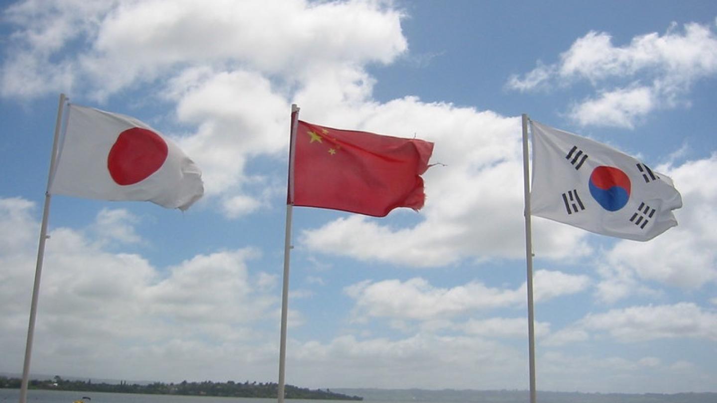 Flags of east Asia on poles near a body of water. Blue sky behind them
